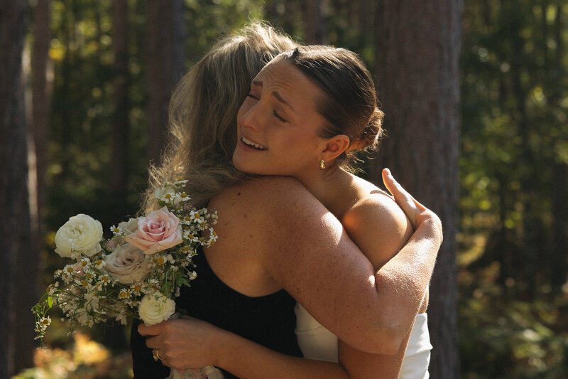 The bride hugs a wedding guest tightly with her white and pink flower bouquet in her hand. She has an emotional expression on her face as she cries over the excitement of her wedding day. They are surrounded by Northern Minnestoa tall pine trees. 