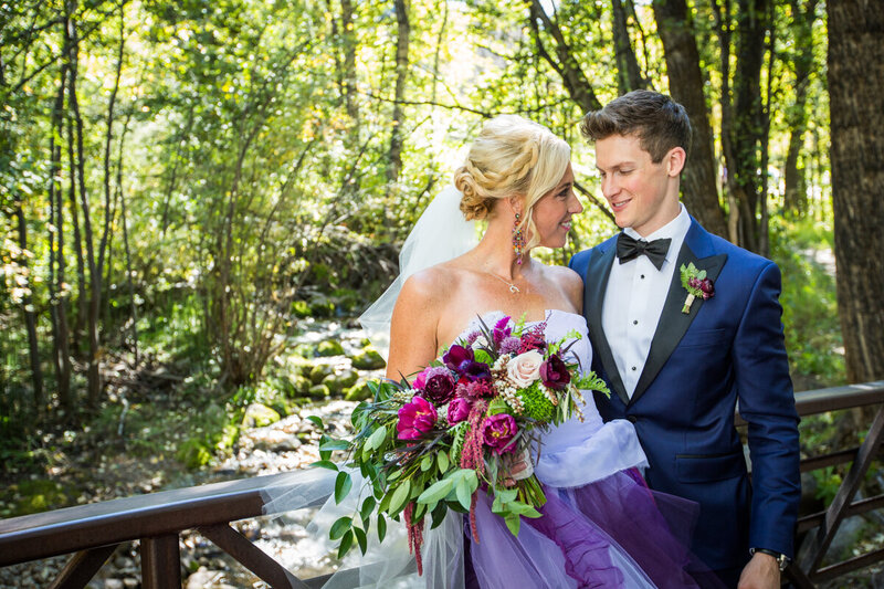 A bride and groom smile at each other leaning on a bride over a stream. The bride's dress is white and purple.