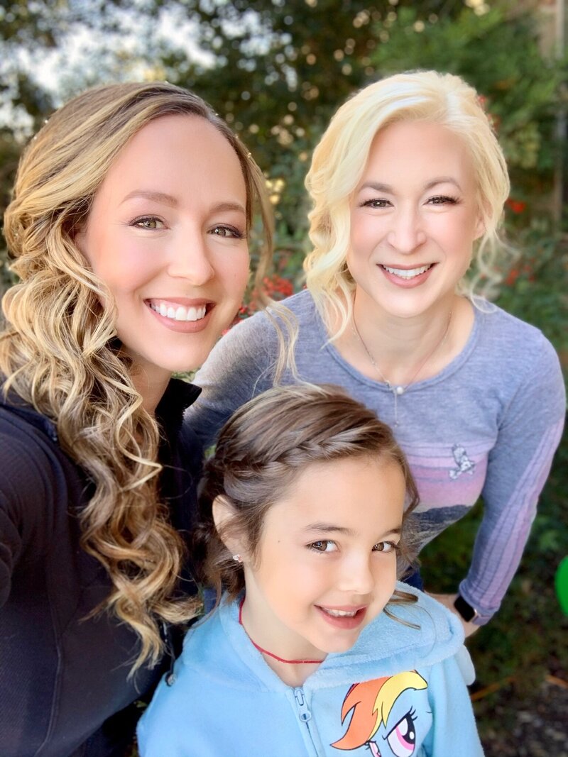 Three people smiling outdoors; two women with wavy hair that could have been styled at a bridal beauty experience in Texas, and a child in a light blue hoodie with a rainbow design.