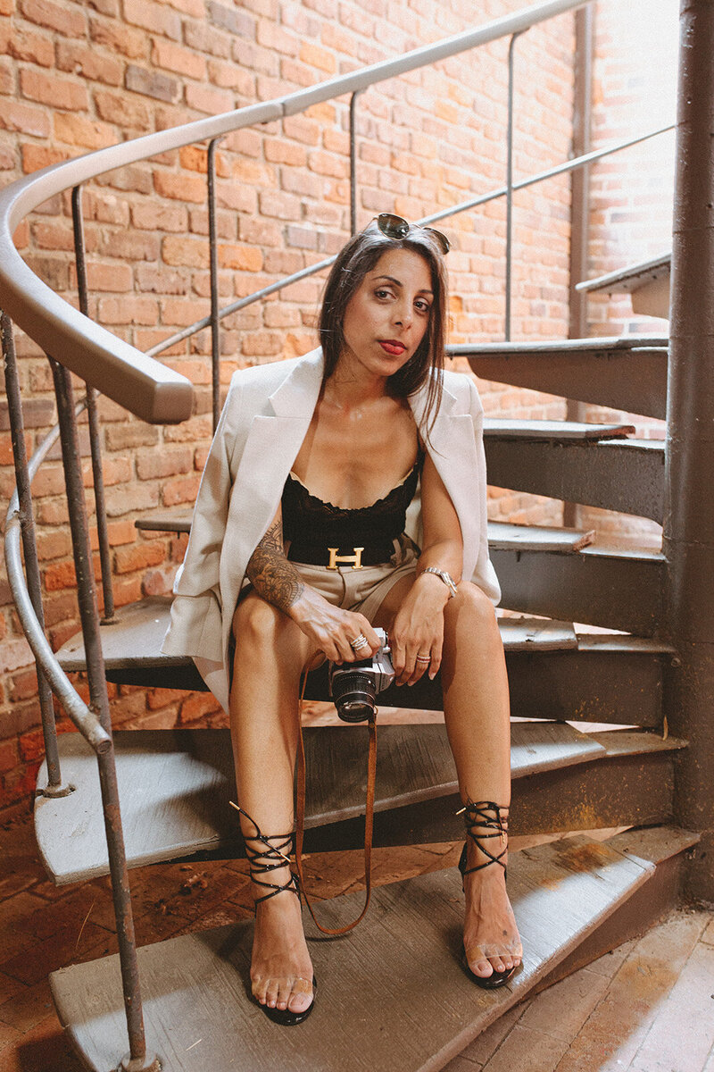 A stylish woman sits on a spiral staircase against a rustic brick wall, holding a film camera. She's dressed in a white blazer, black lace top, designer belt, and strappy black heels, exuding effortless confidence and creative energy.