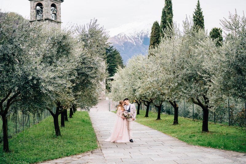newlyweds-walk-olive-grove-against-scene-old-tower-with-bells-lake-como-side-min