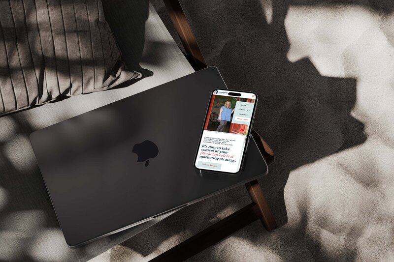 A closed black laptop with an Apple logo sits next to a smartphone displaying a Showit web design for personal brands webpage, both on a cushion in bright sunlight.