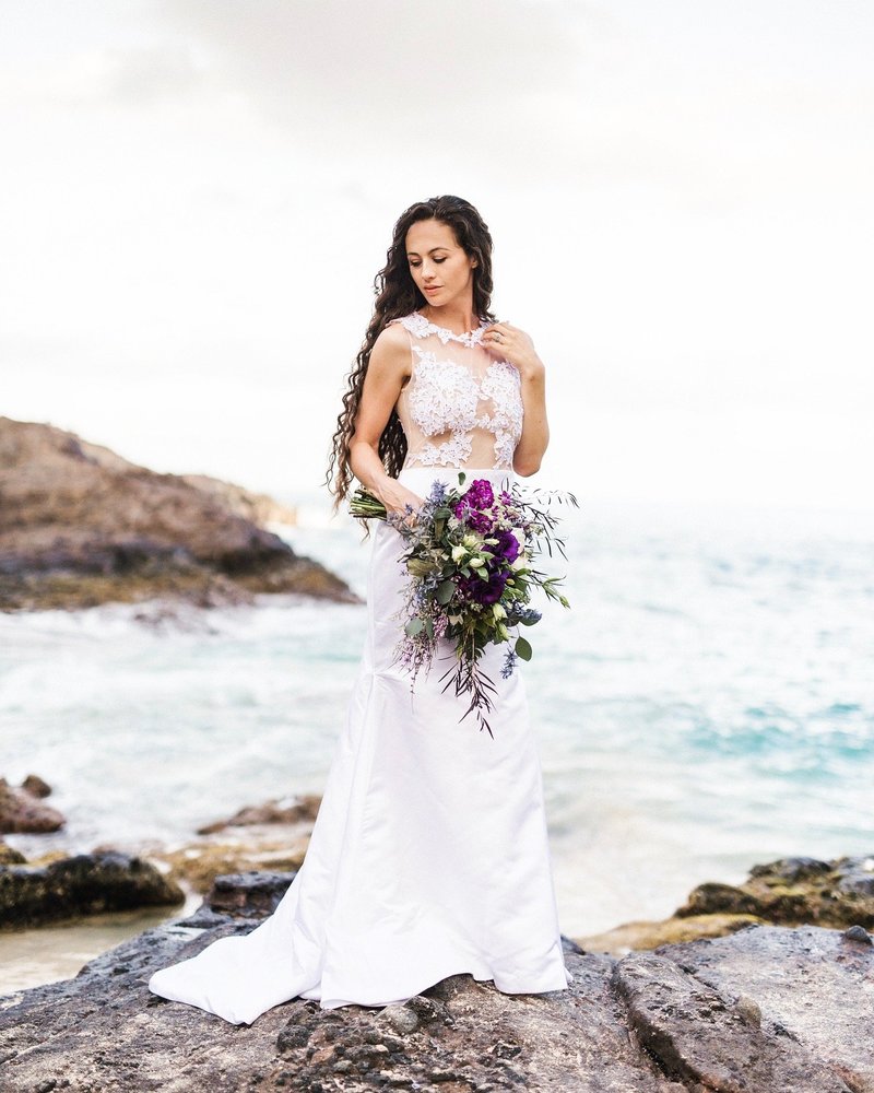 A bride in a lace dress standing by the ocean with long curly brown hair. Wedding photography by Amy Jean.