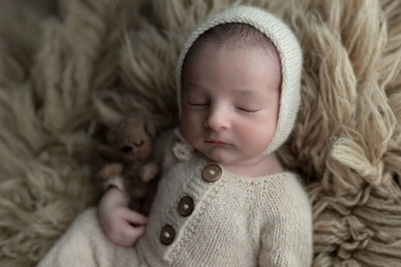 Newborn boy in soft cream knits, peacefully holding a teddy on a textured flokati rug—styled in rustic tones.