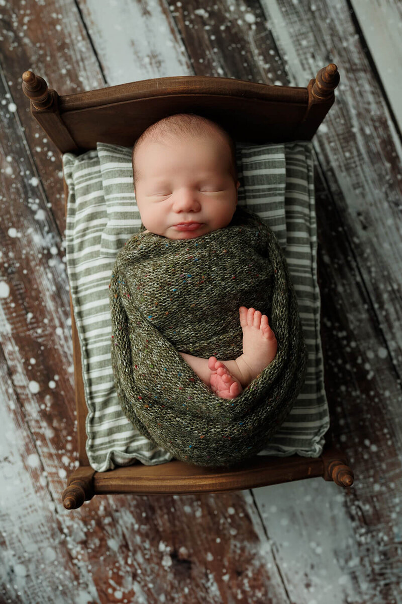 baby boy in a green speckled wrap with his toes out on a tiny bed with coordinating sheet