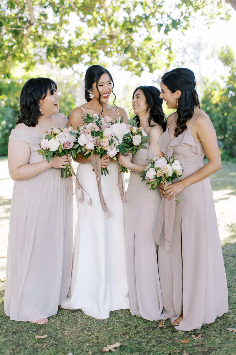 1-radiant-love-events-3-bridesmaids-smiling-at-bride-holding-bouquetss-full-body-outside-greenery-backdrop-romantic-elegant-timeless