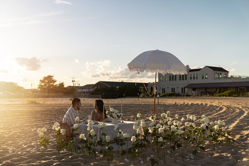 Couple 's sweet moment during a romantic beach picnic with white roses, a fringe umbrella, and ocean views, styled by Yours Truly, a wedding planner and designer in New Jersey