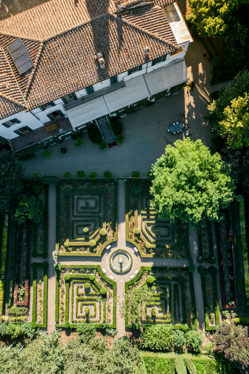 An aerial view of a symmetrical garden with a central fountain, surrounded by greenery and an elegant villa