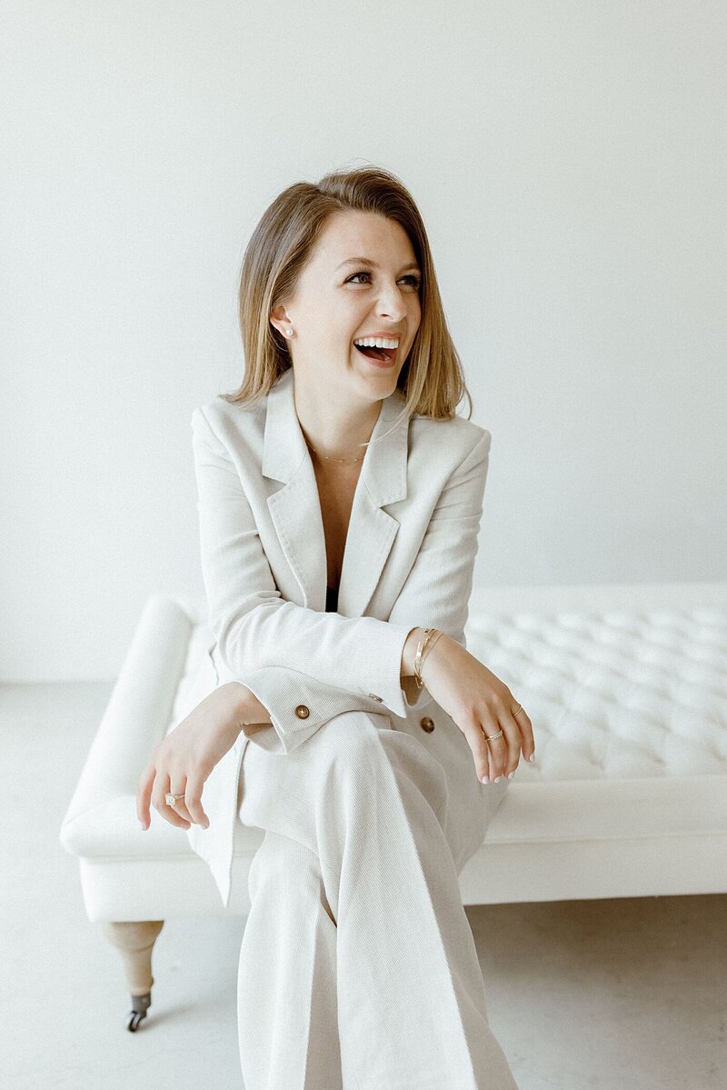 woman in a white pantsuit sitting down and laughing off camera