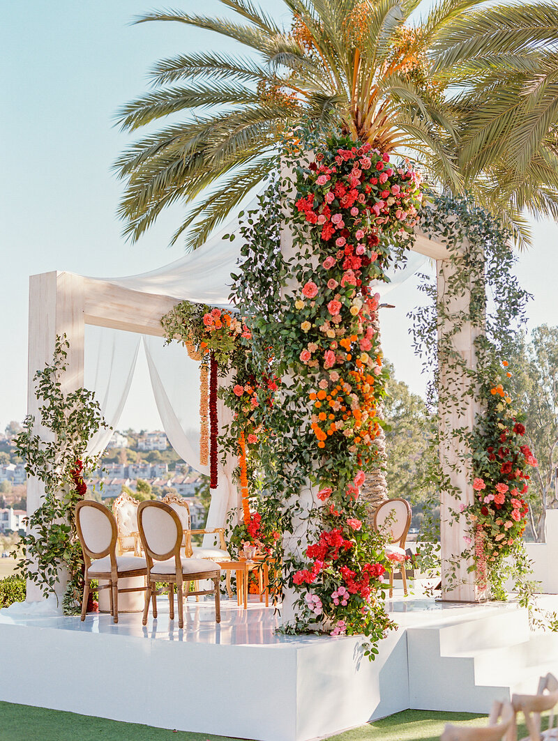 large ceremony stage for indian wedding covered in hotpink, yellow, and orange flowers with white chairs and orange hanging florals
