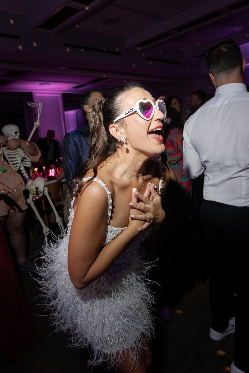 bride in white spaghetti-strap mini dress with sequins and feathers and iridescent heart shaped sunglasses dancing on the dance floor surrounded by guests clutching her chest with her hands  lip singing