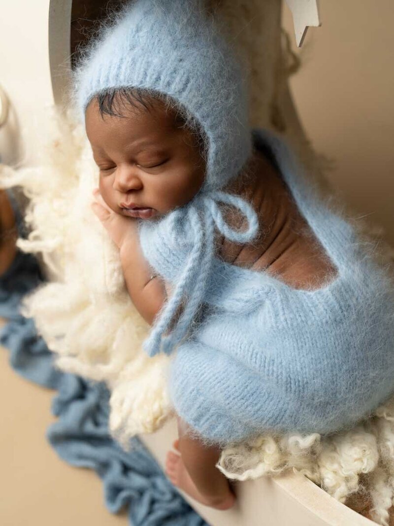 A serene newborn baby sleeps peacefully, dressed in a soft, light blue knitted outfit with a bonnet. The baby is nestled on cozy cream-colored wool.