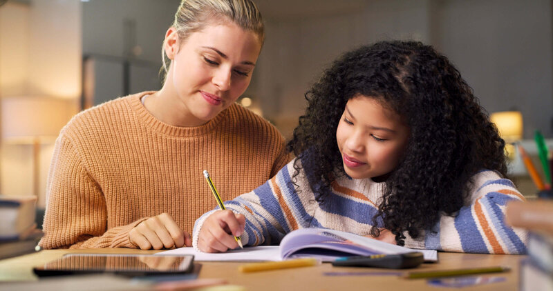 A teacher and a young female student are working on a writing assignment together at a desk
