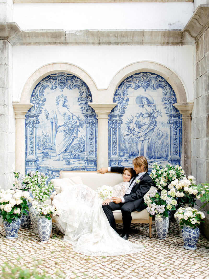 Bride and Groom sit on couch in courtyard of Penha Longa Resort Monastery surrounded by blue Portugese tile and vases of white and green florals