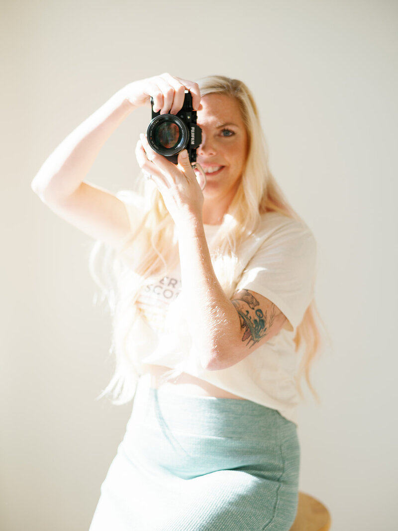 Kristen looking at the camera with her own camera in front of her face on a green background with a blue long skirt and white doc martens, a pink streak in her blonde hair