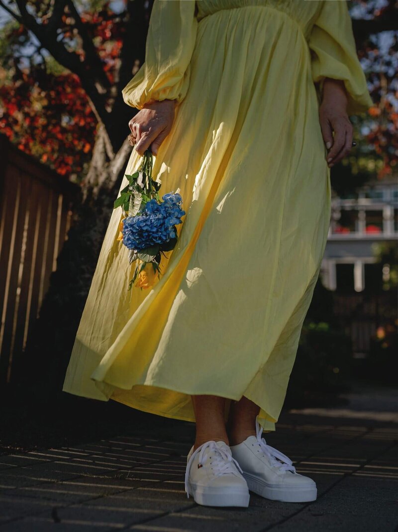 A memoir-worthy scene unfolds as a person in a yellow dress, like an author capturing inspiration, holds blue flowers on a sunlit path with a wooden fence and trees in the background.