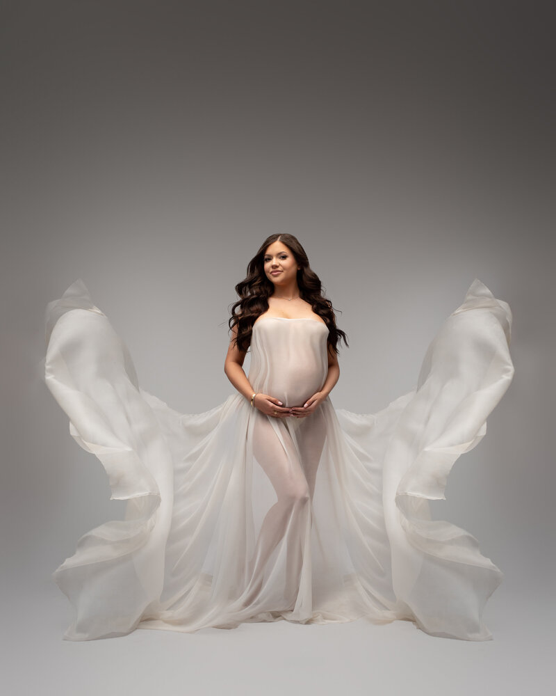 Pregnant mom with glowing skin and long dirty blonde hair wearing long grey tulle and lace maternity dress holding hands on top and bottom of belly on gray backdround
