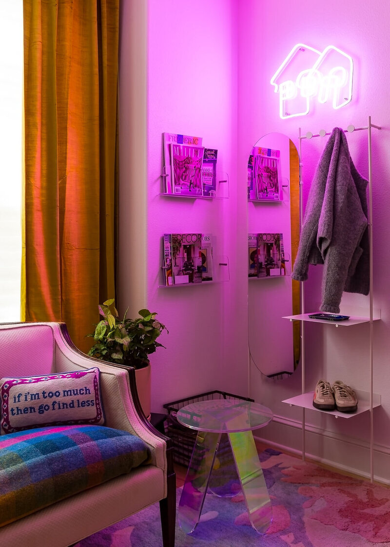 A corner of a room that featuring a neon sign giving off a pink glow, magazine racks hanging on the wall, a colorful rug and the corner of a couch representing colorful home decor.