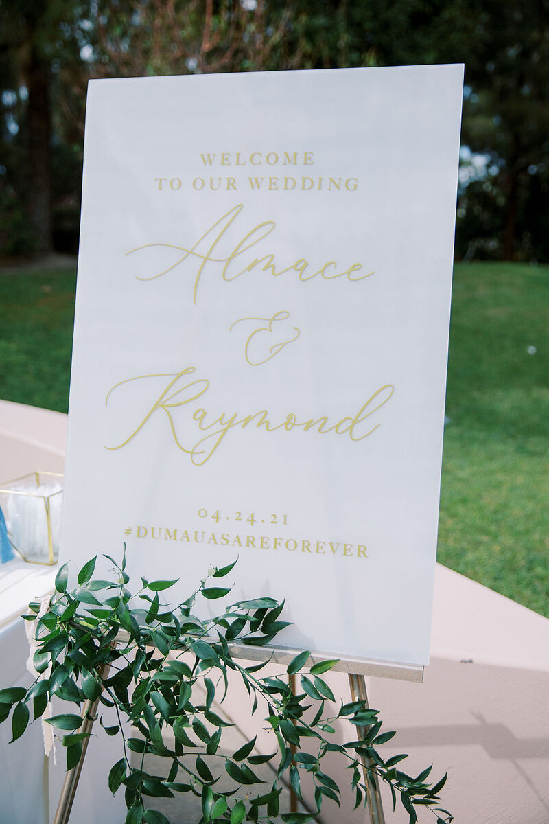 2-radiant-love-events-outdoor-white-welcome-poster-gold-script-on-easell-greenery-below-romantic-elegant-timeless