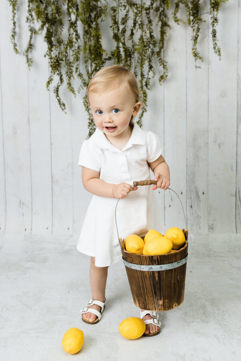 Little girl first birthday summer photo session where she's holding a bucket of lemons