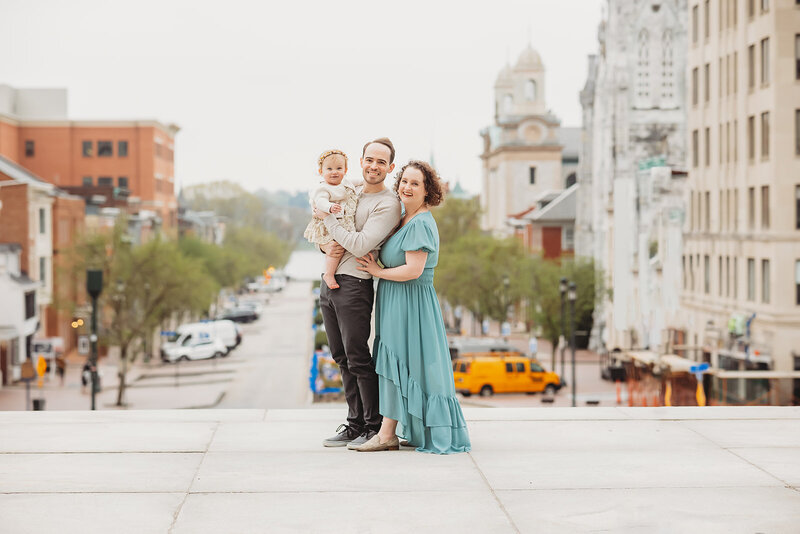 mom, dad, and little girl standing on the steps of the capitol at sunset set smiling for a photo