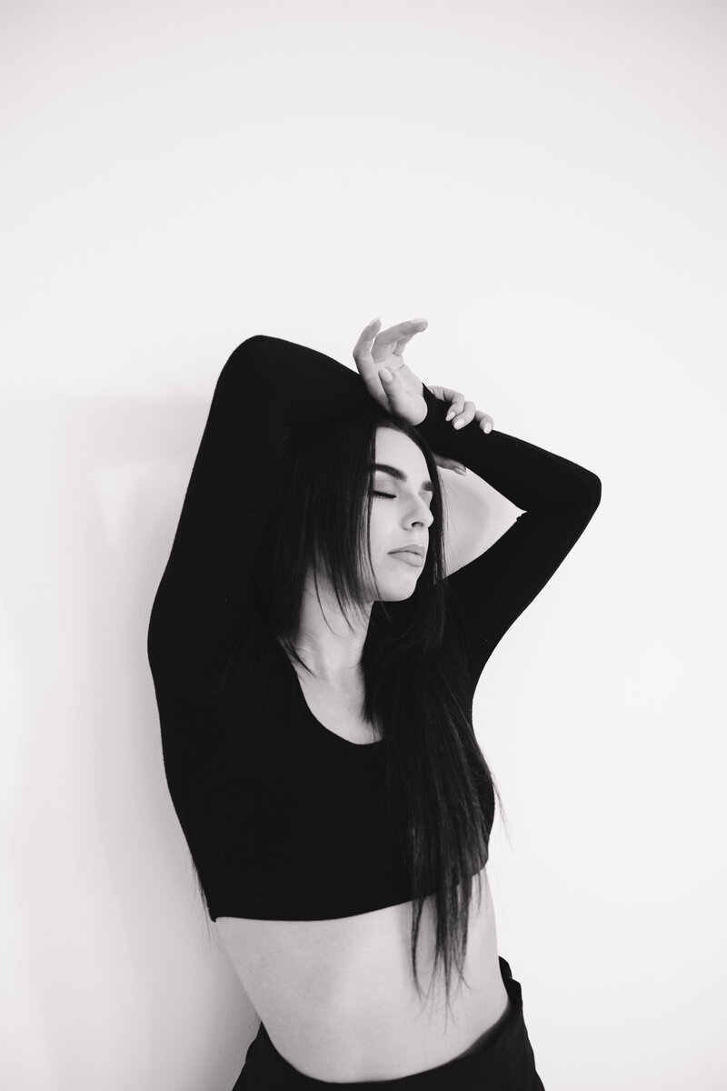 Black and white studio portrait of a woman in a cropped black top with her eyes closed and arms gracefully posed above her head.