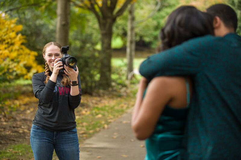 Jen Stitt holding a camera and smiling warmly during an engagement session, showcasing her enthusiastic and approachable style while capturing stunning moments for engaged couples.