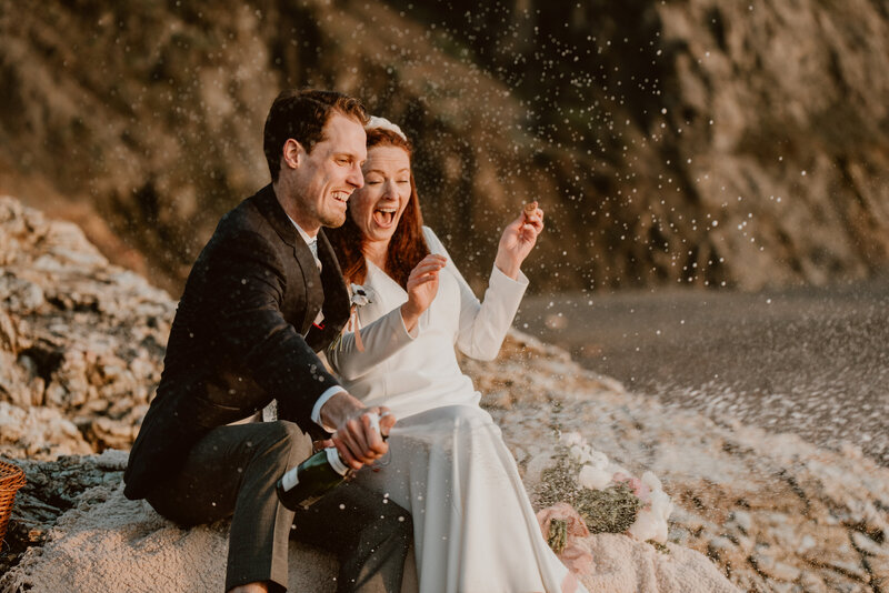 Couple exclaiming in joy as they pop a bottle of champagne on the beach in Sonoma Coast State Park during their wedding day