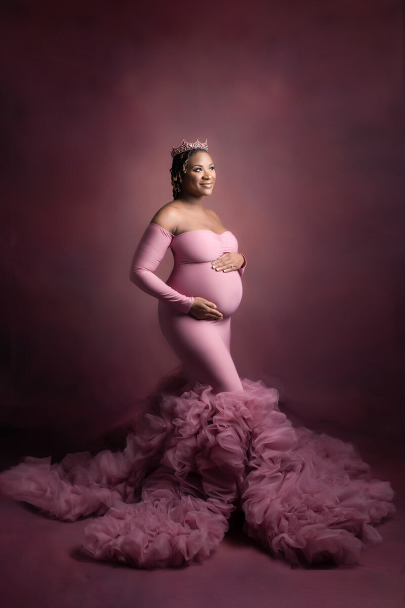 Maternity photography — pregnant woman in dramatic pink mermaid gown and crown, posed against rich burgundy backdrop