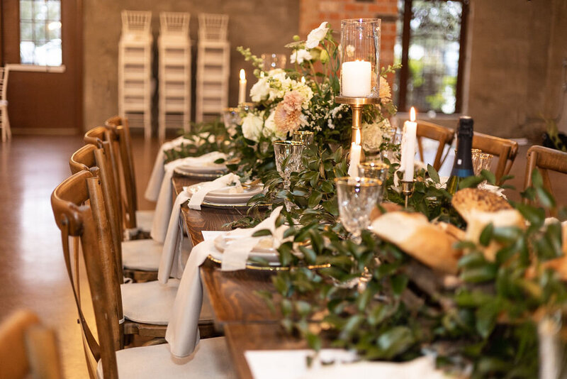 A close-up view of a rectangular wooden farmhouse table decorated with greenery, taper candles, glassware, and layered neutral plates.