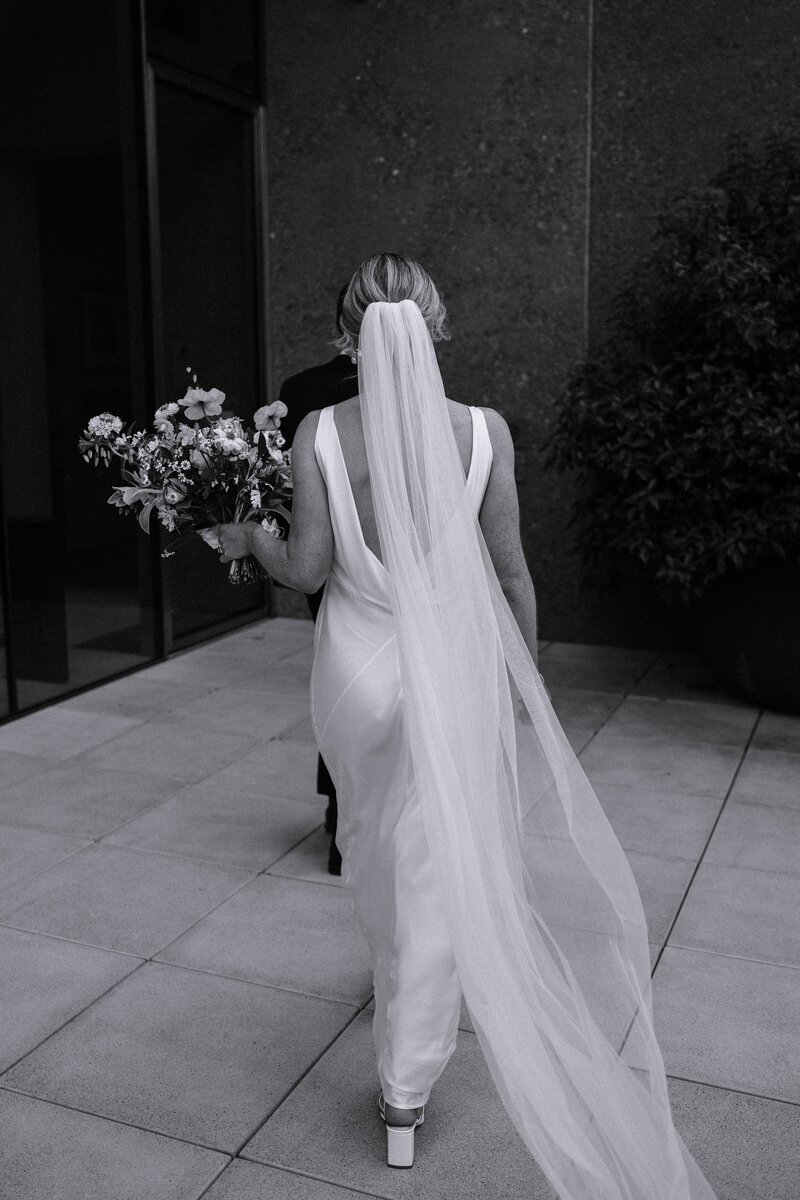 a black and white shot of the back of a bride as she walks towards the concrete wall of ranvenscar house museum in christchurch