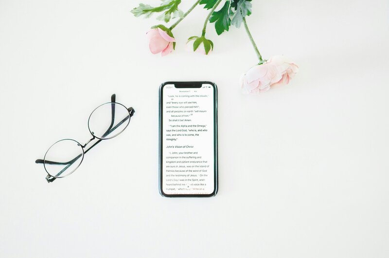 A phone lying on a counter with bible verse on the screen. There is a light pink long stemmed rose framing the picture and a pair of glasses to the left of the phone.