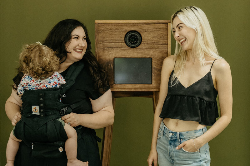Clarae and Taressa, wedding and event planners in Oregon, smile at each other in front of their open air photo booth and Taressa has her child on her chest facing away from the camera.