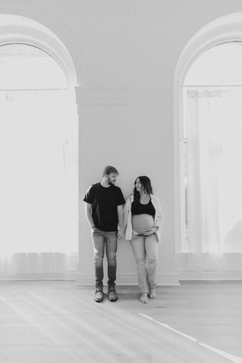 Black and white maternity photo of a couple in a natural light studio, holding hands and looking at each other, taken during a lifestyle maternity session in the Midwest by a Michigan and Ohio maternity photographer.