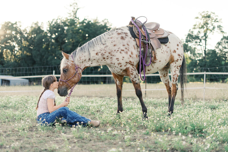 A girl sitting in a field with her horse, giving her horse a kiss