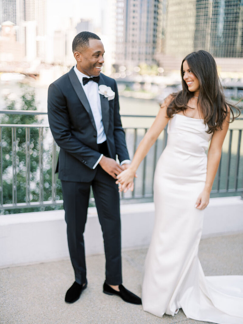 A joyful couple holding hands on a bridge over the Chicago River, the bride in a sleek white dress and the groom in a classic black tuxedo, with the city skyline in the background.