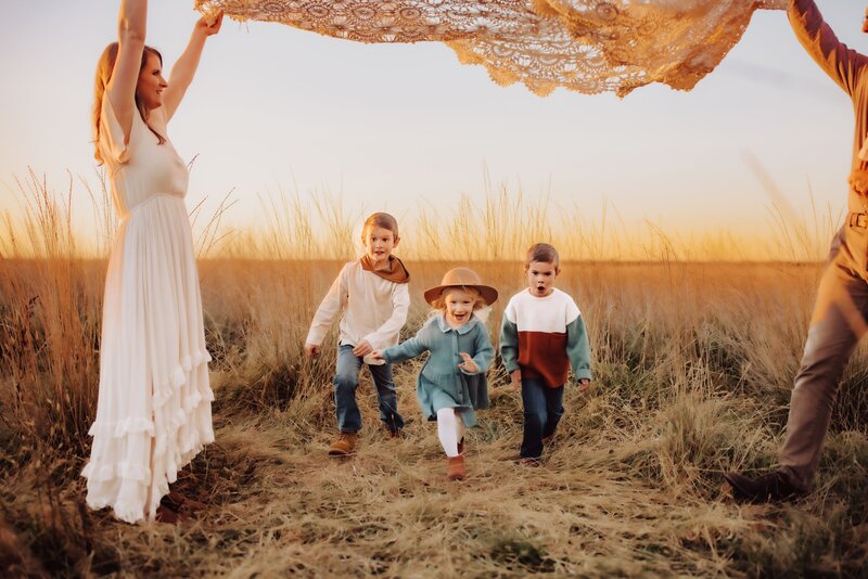 A mother and father and their children outside. The mother is dressed in a white wedding dress and they are holding their veil in the air while the children run beneath it.