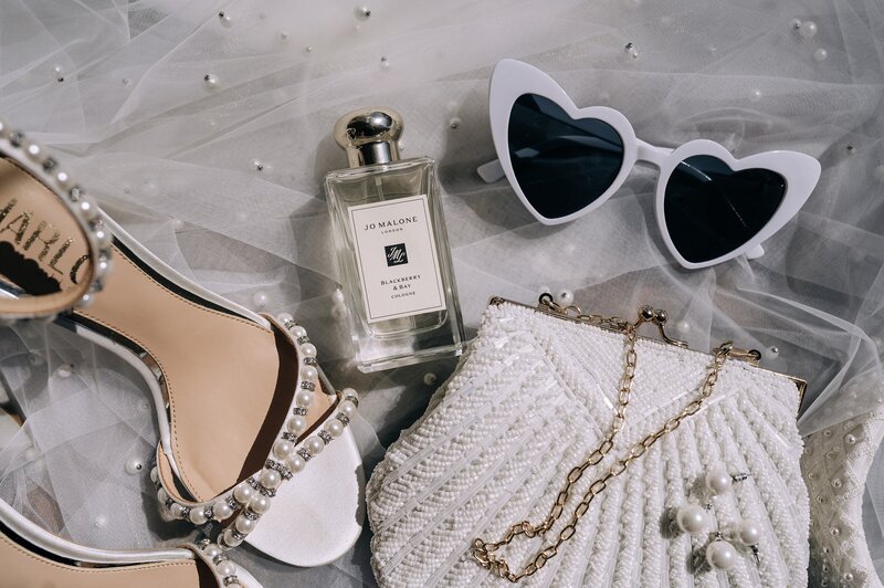 detailed photo of harlo pearl shoes and jo malone perfume with white sunglasses and beaded white clutch at a christchurch wedding photo