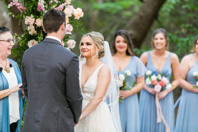 Emotional ceremony moment with the bride in a tulle gown and groom in a grey suit, surrounded by pastel blooms—pure romance.