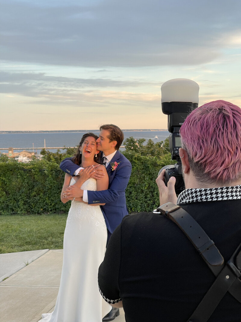 A bride and groom hugging while a photographer takes a picture of them.