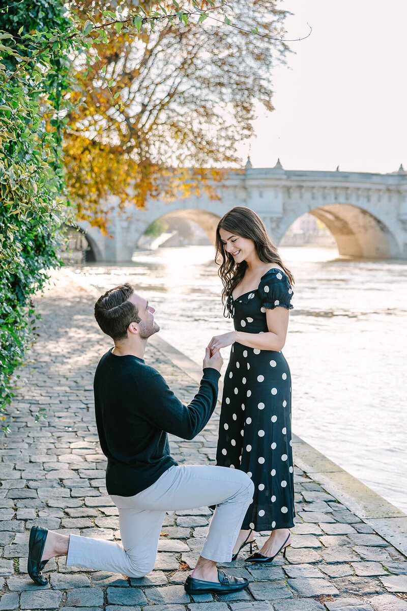 Man proposing to smiling woman in a polka dot dress as she reaches out her hand, captured on Île de la Cité near Pont Neuf during a romantic Paris proposal photoshoot.