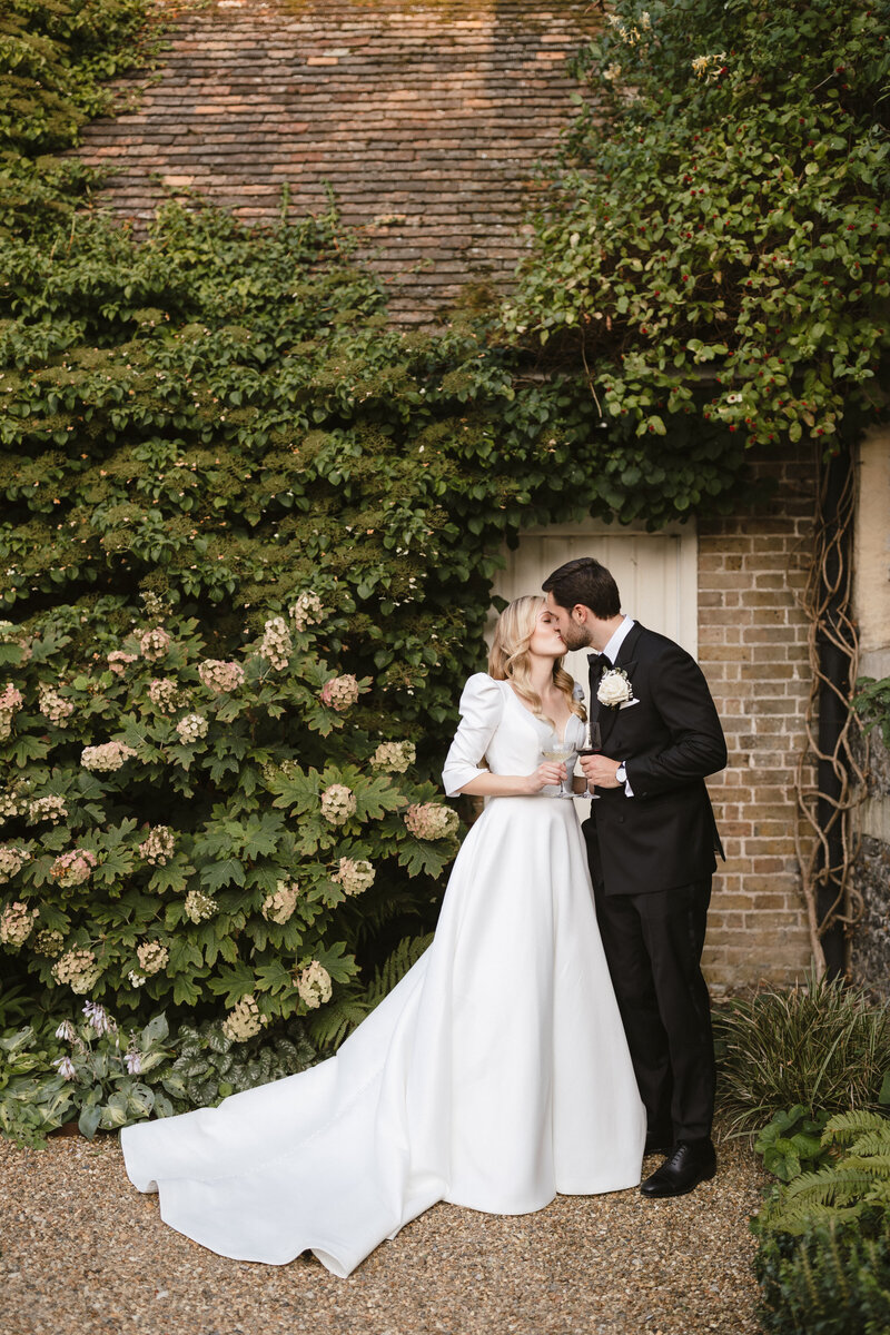 A couple, dressed in a wedding gown and suit, sit happily on the back of a vintage convertible at The Farmhouse at Redcoats. The bride holds up a bouquet as they pose in a gravel courtyard with rustic buildings in the background. The black-and-white image captures their timeless joy.