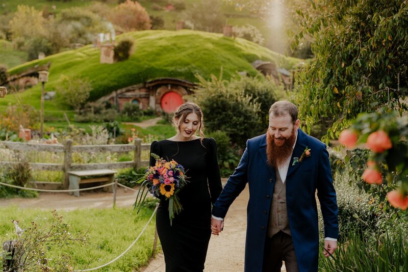 A bride wearing a black dress with a groom in a blue suit walking in the gardens at Hobbiton in the Waikato while photographed for their wedding photoshoot