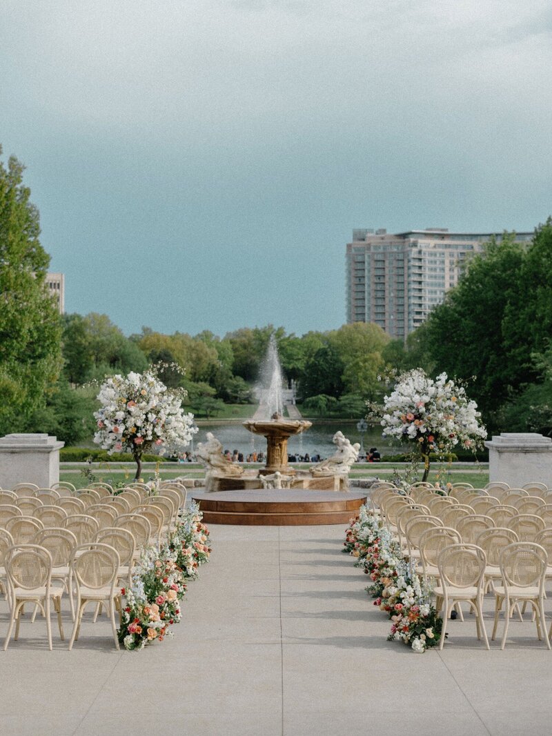 Vintage-style chairs with flowers on either side of a concrete aise leading to a park fountain and blossoming trees in a park with a high-rise building in the background