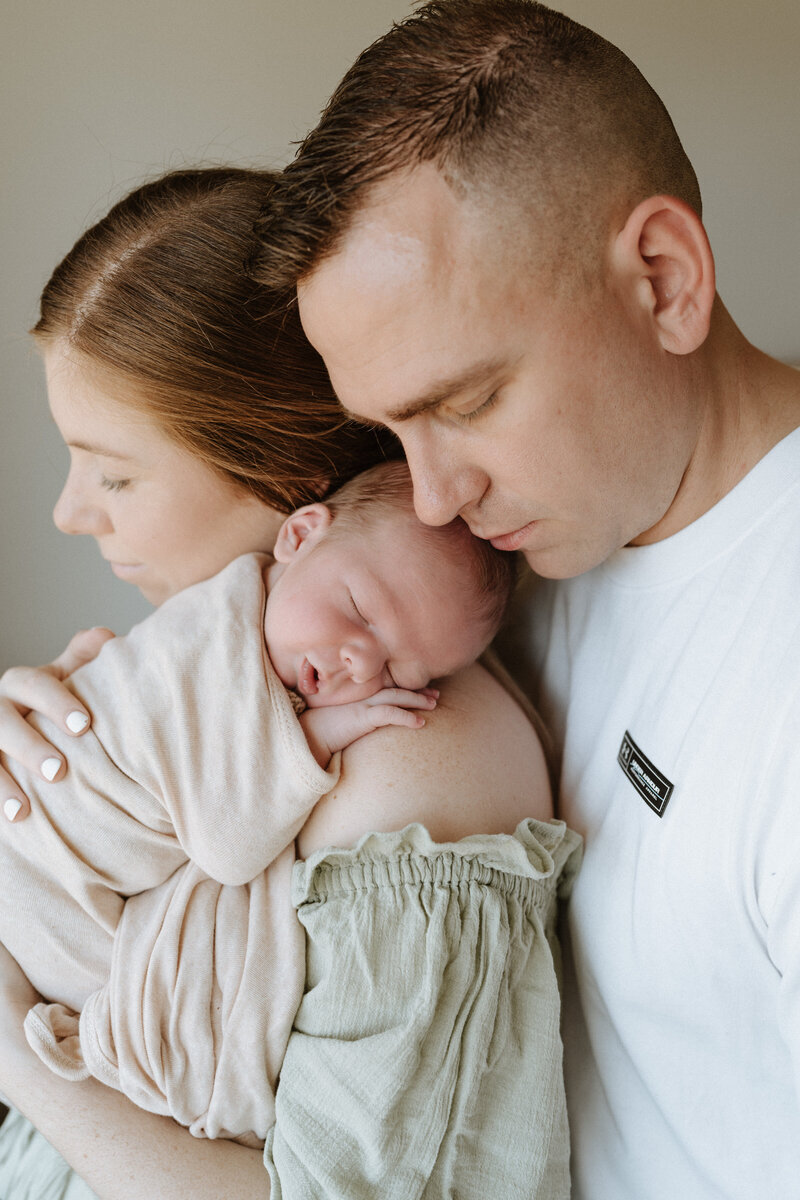 Mother cradles sleeping newborn on her shoulder while the father gently rests his head against the newborn during an in-home newborn photoshoot in Brisbane.