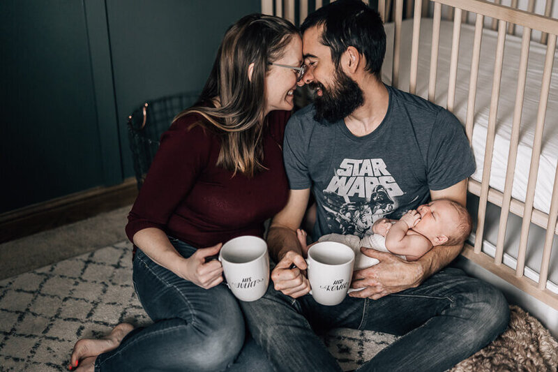 A father hugs his wife and newborn baby from behind, as mama gazes down at her baby wearing a cloth diaper.