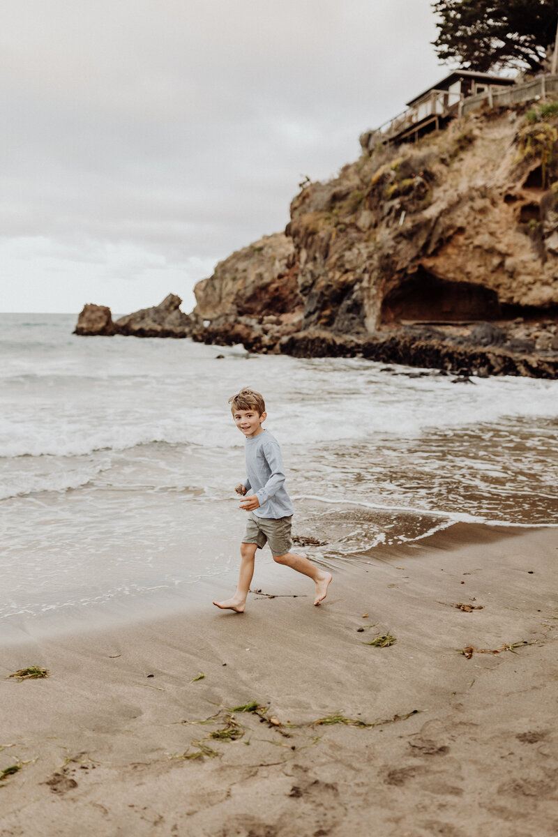 A young boy running along the beach with the rocks and water behind him during a candid family photoshoot with Christchurch photographer Lucy Jane Photography