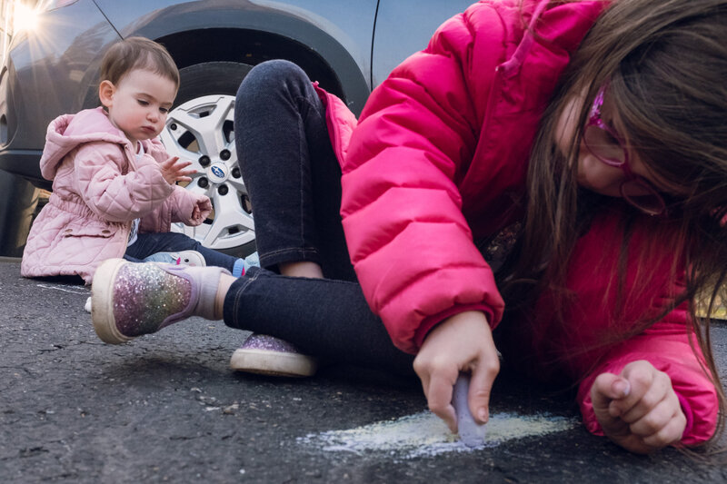 Two little girls playing with chalk on the ground in front of a car.
