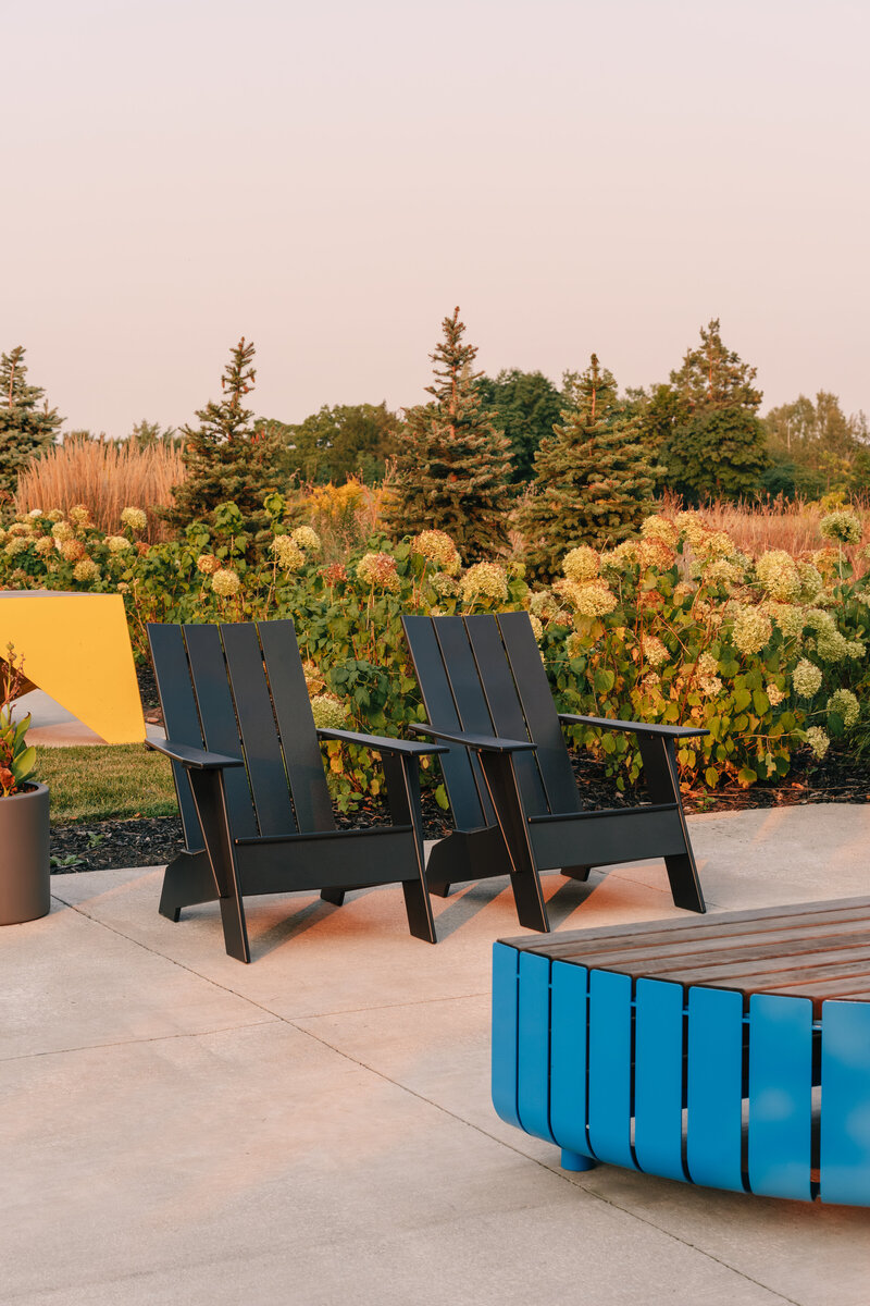 Two comfortable black Muskoka chairs and a blue wood paneled coffee table on a concrete patio, positioned in front of lush flowery bushes and trees