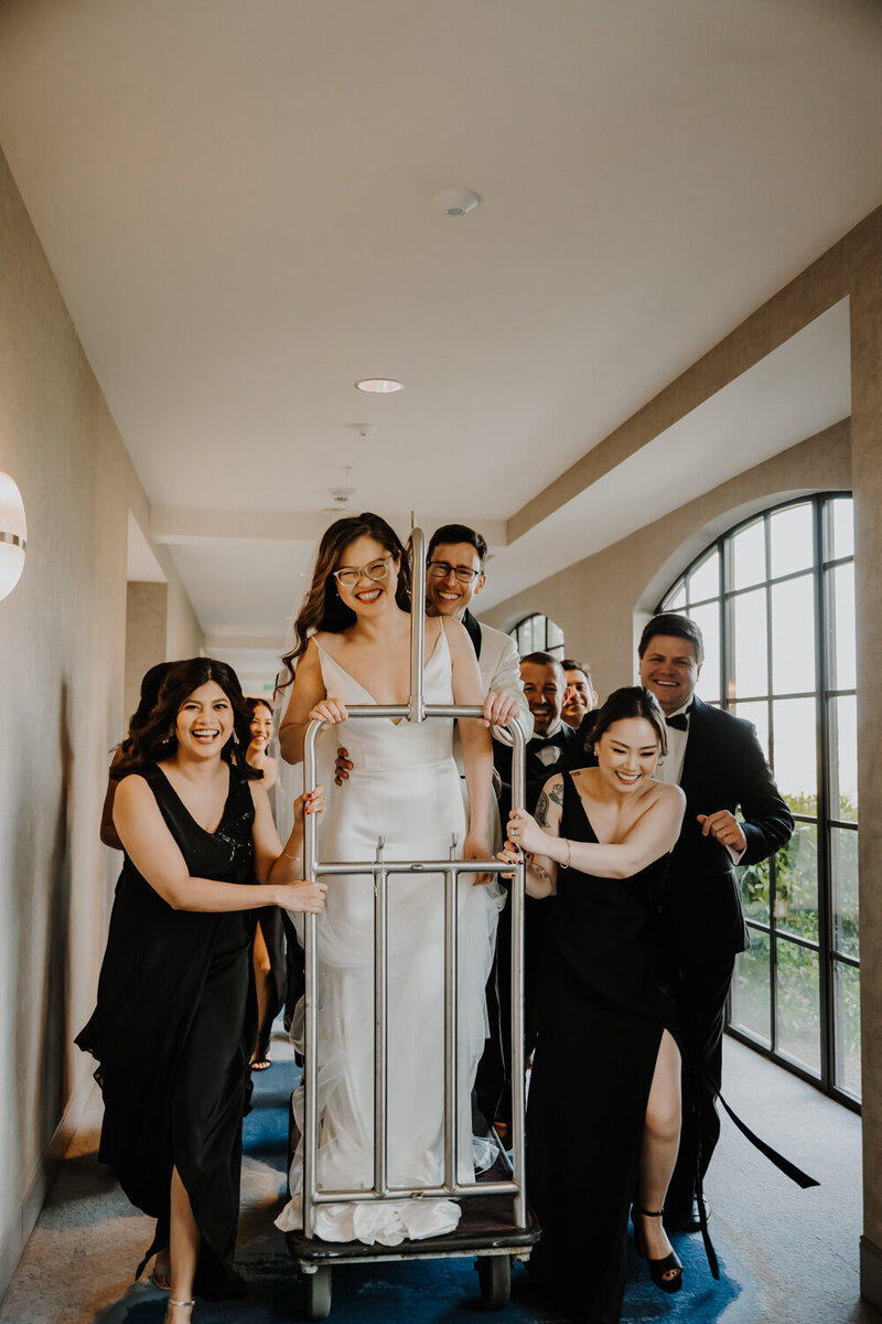 bridal party pushing bride on luggage cart while laughing